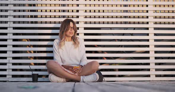 Young Woman Listens to Music From Her Smartphone While Sitting on White Wooden Pier alt