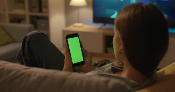 Young woman sitting on the couch at home and using her touch screen smartphone, green screen mock-up