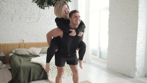 Man Doing Squat Exercise Hilding Woman on His Back Instead of a Barbell at Home