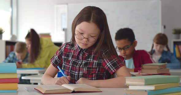 Smart Schoolgirl Sitting in Classroom and Writing in Textbook alt