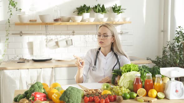 An Attractive Female Nutritionist is Sitting at a Table with a Lot of Fresh Vegetables and Fruits alt
