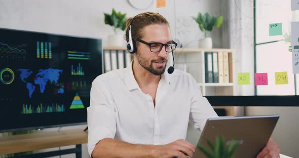 Man Sit at Workplace in Headset Looking at Laptop During Talking to Customer Using Remote Web Chat alt