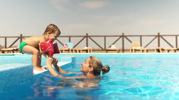 Mom in a Bikini Helps Her Son in Oversleeves to Jump From the Side to the Pool with Clear Water alt