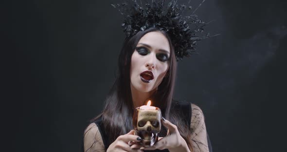 Mystical Female in Black Attire Holds a Skull with a Burning Candle and Looking at the Camera alt