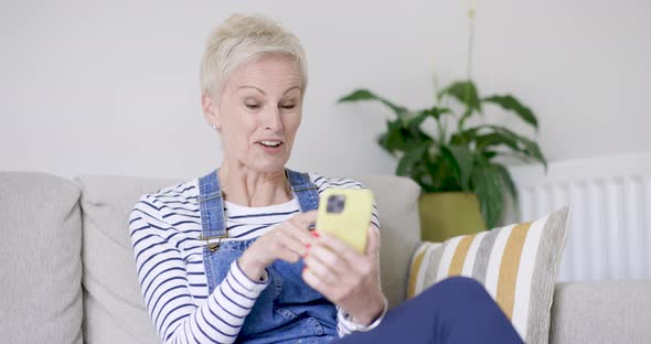 Mature woman sitting on sofa making watching video on smartphone