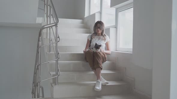Pretty Woman Strokes Colorful Stray Cat While Sitting on White Steps of Staircase of Apartment alt