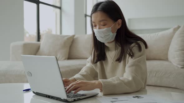 A Thoughtful Asian Woman in a Medical Mask Using a Laptop is Working in the Living Room alt