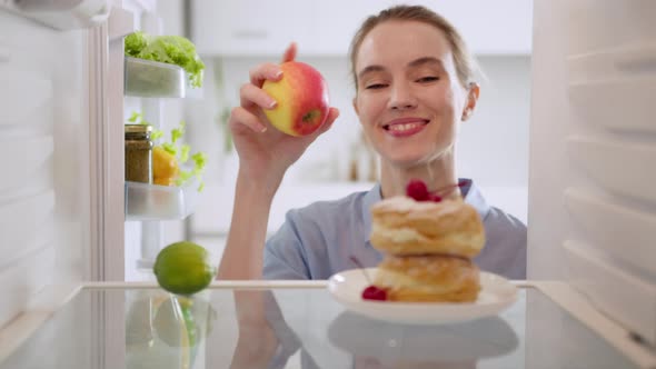 Cheerful woman taking apple instead of cake from refrigerator.