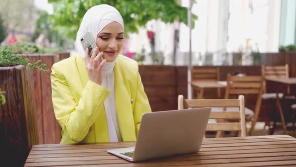 Smiling Young Muslim Woman in Headscarf Talking on the Phone and Using Laptop in Cafe