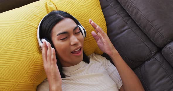 Gender fluid male wearing headphones enjoying listening to music while lying on the couch at home alt