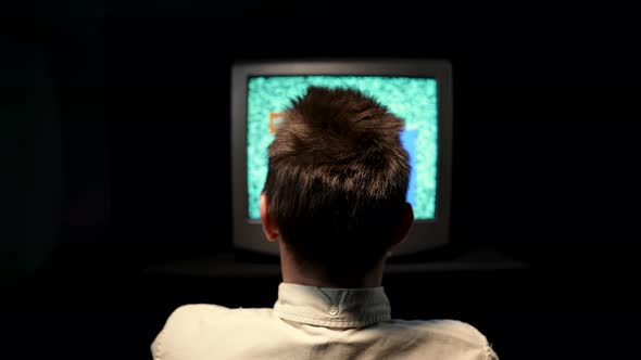 Rear View of a Man Sitting in Front of an Old TV in a Dark Studio on a Black Background alt