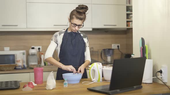 Woman Cooking Muffins Whilst Looking the Recipe at Laptop in the Kitchen at Home alt