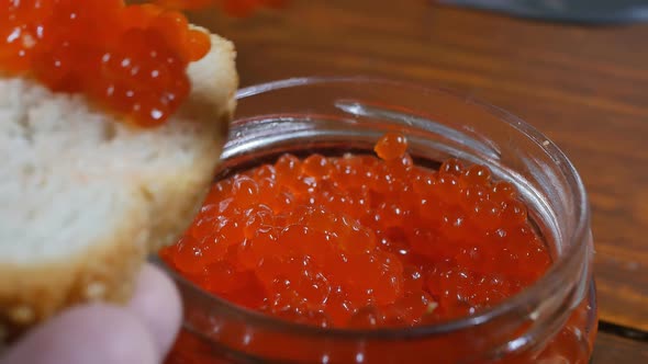 a Man's Hands Lay Red Caviar on a Piece of Bread From a Glass Jar Using a Stainless Steel Table
