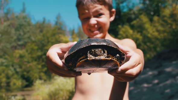 Boy Holds Turtle in Arms and Smiles Viciously on River with Green Vegetation alt