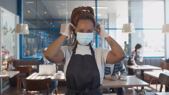 African Waitress Wearing Protective Mask and Gloves Standing in Cafe During Covid-19 Preventing. alt
