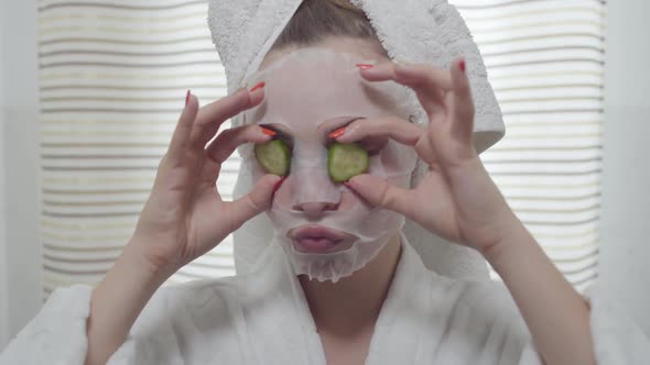 Attractive Young Woman Eating Cucumber with the Sheet Mask on Her Face in the Bathroom alt