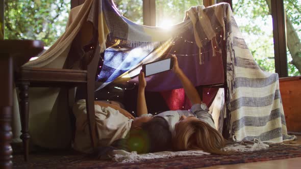 Asian mother and daughter using digital tablet while lying under blanket fort at home alt