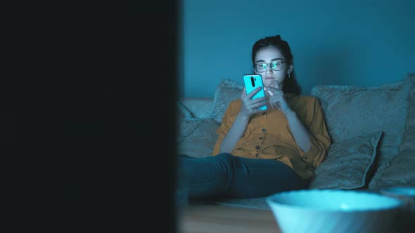 Alone Teen Girl Sits in the Evening Near Tv and Uses the Phone Chating with Friends and Smiling