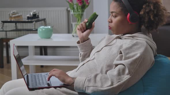 Side View of African American Serious Woman Eating Cucumber Typing on Laptop Keyboard Sitting on Bag alt