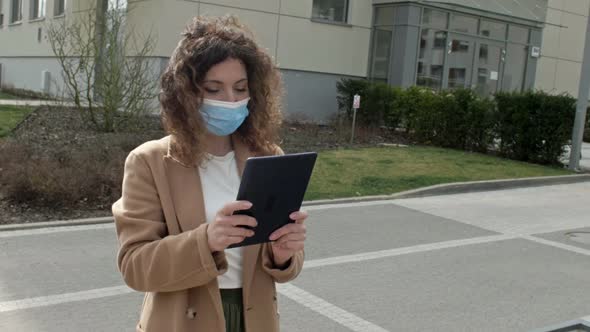 Beautiful Woman in a Protective Mask with a Tablet in Hand Walks Along a Deserted City Street alt