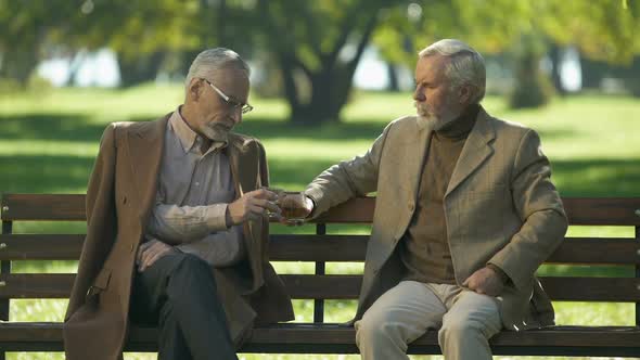 Two Upset Senior Gentlemen Drinking Whiskey on Bench in Park, Unhappy Memories alt
