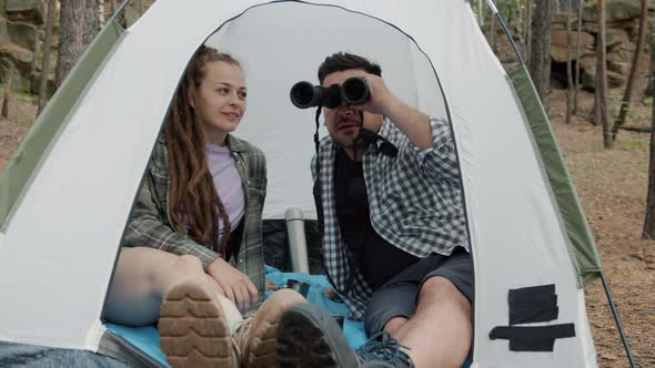 Young Happy Man and Woman Talking and Looking at Forest Through Binoculars Inside Tent on Autumn Day alt