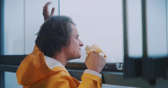 Young man looking out of foggy window while smiling and eating a banana alt
