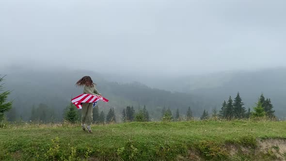 happy little girl is running and jumping carefree with open arms in cloudy mountains. Holding USA 