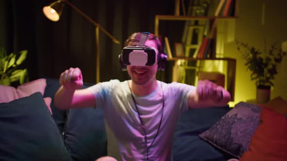 Young Man Wearing Virtual Reality Glasses and Headphones Playing Video Games Gamer Sitting on Sofa alt