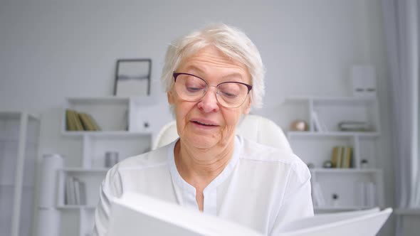 Old corporate manager in glasses reads white book sitting at online video conference alt