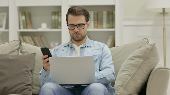 Young Man with Smartphone Working on Laptop at Home alt
