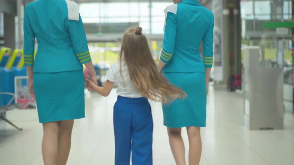 Back View of Happy Caucasian Girl Holding Hands of Professional Stewardesses and Walking To Airport alt