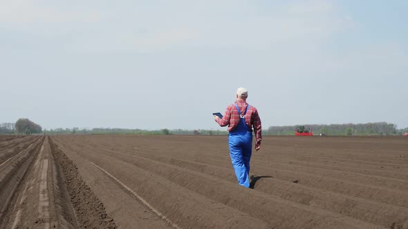 Farmer, Agronomist Walks on Field with Special Soil Rows. He Tests, Using Tablet, Quality of Potatoe alt