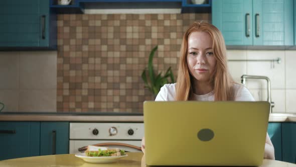 young freelance woman eats sandwich and works on  laptop at home in the kitchen alt