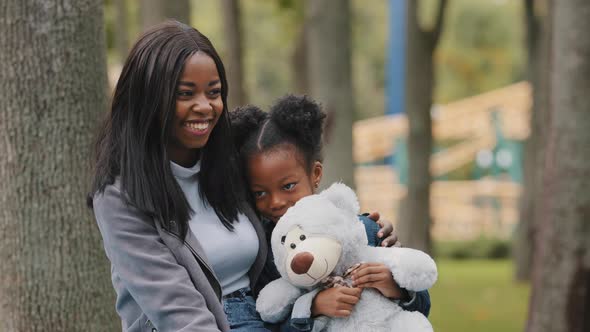 Joyful Mom and Daughter Sitting on Bench in Park Young Mother Hugs Child Shy Little Girl Holding alt
