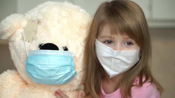 Lttle Girl Sits with Teddy Bear in Protective Masks at Home and Looks at Camera