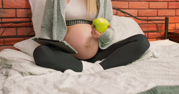 Pretty Pregnant Woman Having Rest on the Bed Among Pillows Biting and Eating a Green alt