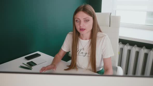 Caucasian Young Woman Sitting at Desk in Office and Gesticulating While Communicating with Another