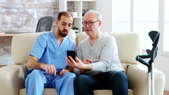 Male Nurse Helps a Retired Old Man To Use a Smartphone alt