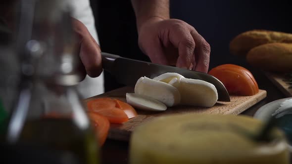 Chef Cuts Mozzarella with Knife on Board. Tomatoes, Olive Oil, Cheese on Table