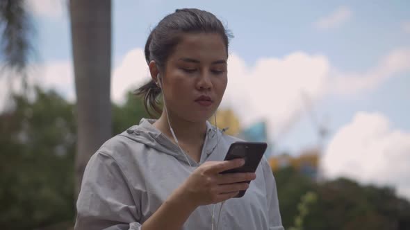 Asian woman fitness runner using mobile phone listening music at a public park.