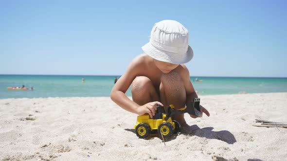 On a Sunny Summer Day, a Child Plays with a Toy Excavator on a Sandy Beach, with an Azure Sea  alt
