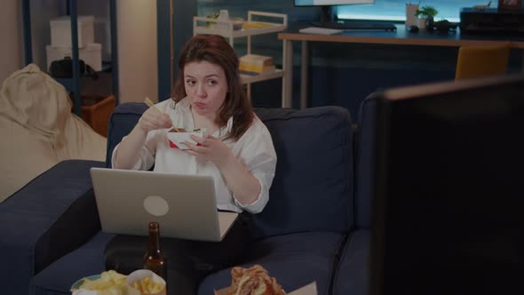 Young Woman Using Laptop on Sofa While Eating Asian Food alt