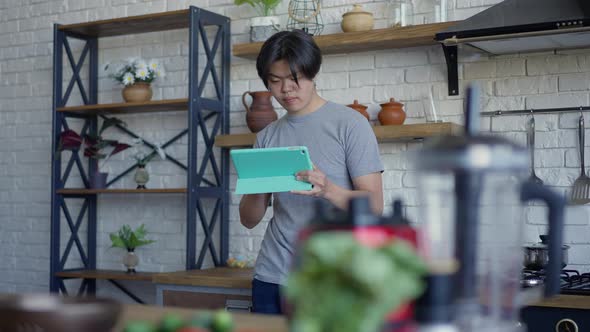 Portrait of Young Thoughtful Man Standing in Kitchen Surfing Internet on Tablet alt