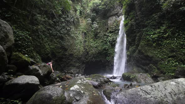 Male Backpacker Tourist Walking at the Foot of a Massive Waterfall. alt