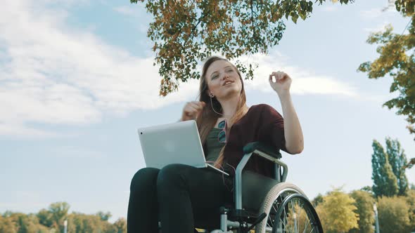 Happy Caucasian Woman in the Wheelchair Listening the Music on Laptop Under the Tree. Low Angle alt
