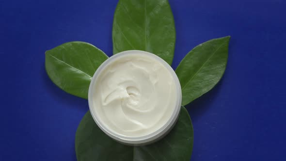 Water Flowing Slow Mo Around Open Jar of Beauty Cream on Green Leaves on Blue