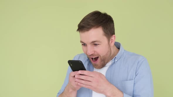 Portrait of Happy Young Guy Enjoying Success on Mobile Phone in Studio on Olive Background alt