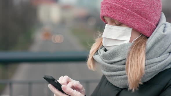 COVID-19 Coronavirus Pandemic. Woman in Mask and Gloves Uses Smartphone Outside