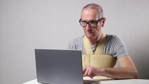 Senior Man with Fixating Bandage Works on Laptop at Desk alt